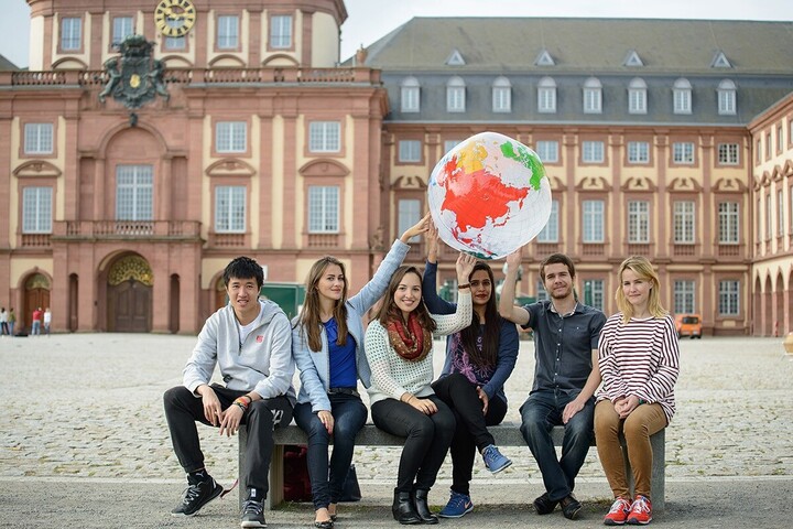Six students sit in front of the University of Mannheim. Some of them are holding a globe above their heads.