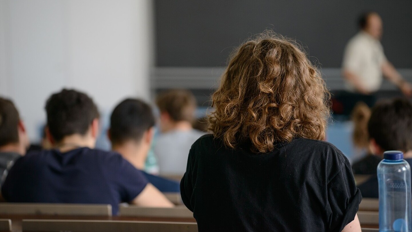 Shot from behind in a lecture hall. The focus is on a female student with brown curly hair and a black top. Other students are sitting in the background.