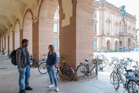 A student and a female student are standing in a corridor that borders the “Ehrenhof” on the left and talking. There are many bicycles in the arches of the corridor.