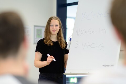 A student is standing at a flipchart. She is holding an open Sharpie in her hand and looking towards the audience. Two heads can be recognized on either side of the picture.