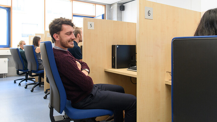 Aufnahme aus dem mLab. Einige Studierende befinden sich in dem Labor. Ein Student lehnt sich zu dem Arbeitsplatz einer Studentin rechts neben ihm.