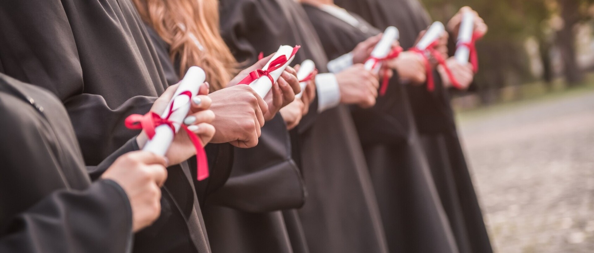 Graduates in black robes. In their hands they hold a white roll of paper with a red ribbon.