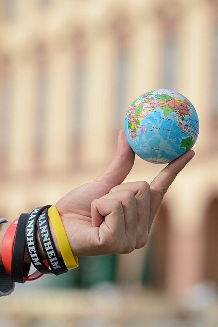 A small globe is held with two fingers. Black, yellow and red bracelets are worn on the wrist, with ‘Mannheim’ written in white on the black bracelets. The University of Mannheim can be recognized in the background.