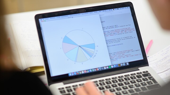 Blick auf einen aufgeklappten Laptop. Auf dem Bildschirm befindet sich linksseitig ein Kreisdiagramm, rechtsseitig und m Hintergrund der Code für das Diagramm. Zwei Personen schauen auf den Laptop.